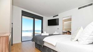 A modern bedroom with a large bed, a flat-screen TV, a bathroom with a mirror, and a view of the ocean through a sliding door. - Villa Oasis - Five Bedroom Villa with Private Pool (Trogir)