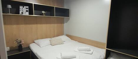 Een hotelkamer met een bed, een nachtkastje met lamp, een plank met decoratieve spullen, en een kledingkast.