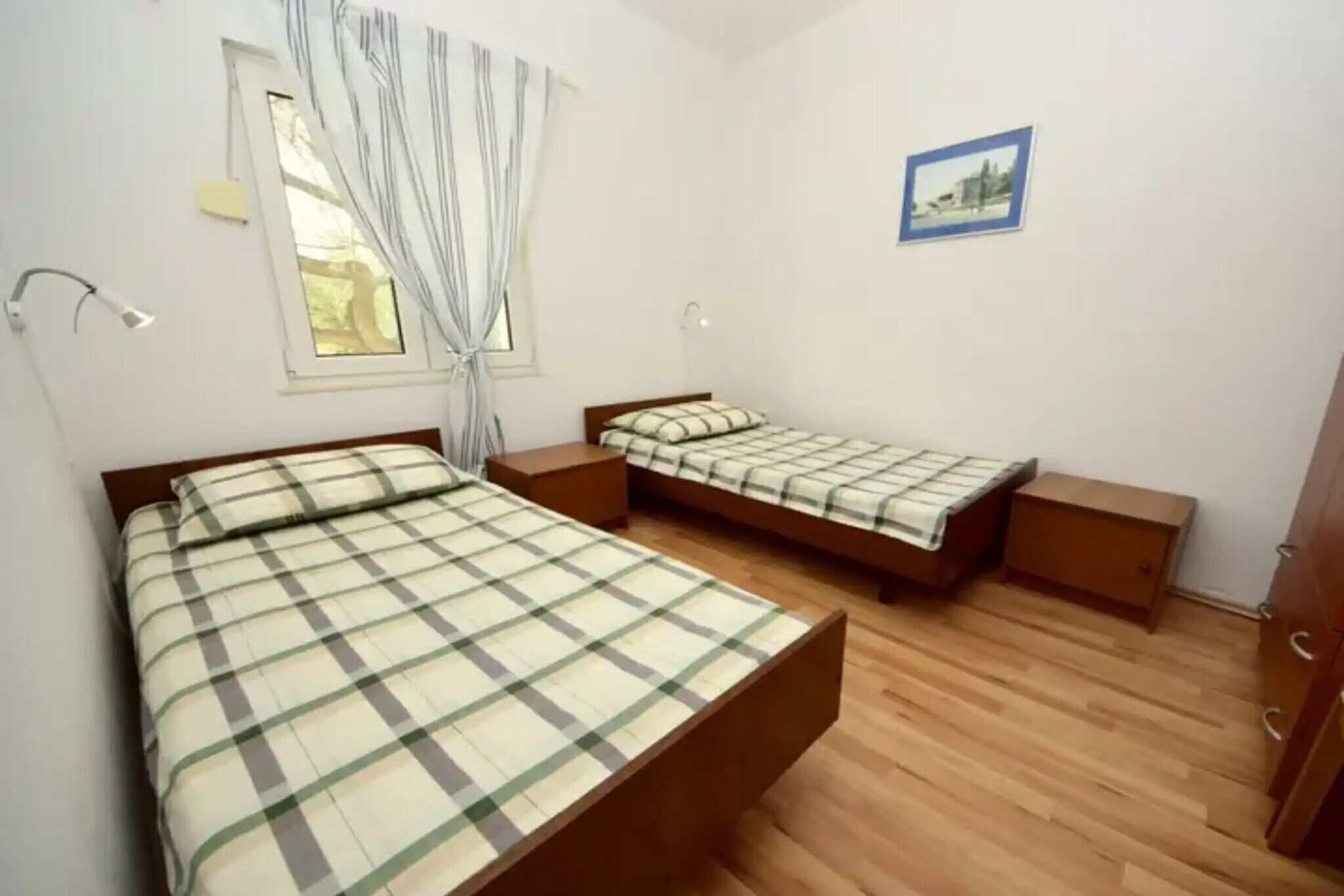 A bedroom with two beds, wooden flooring, a window with curtains, a bedside lamp, and a framed picture on the wall.