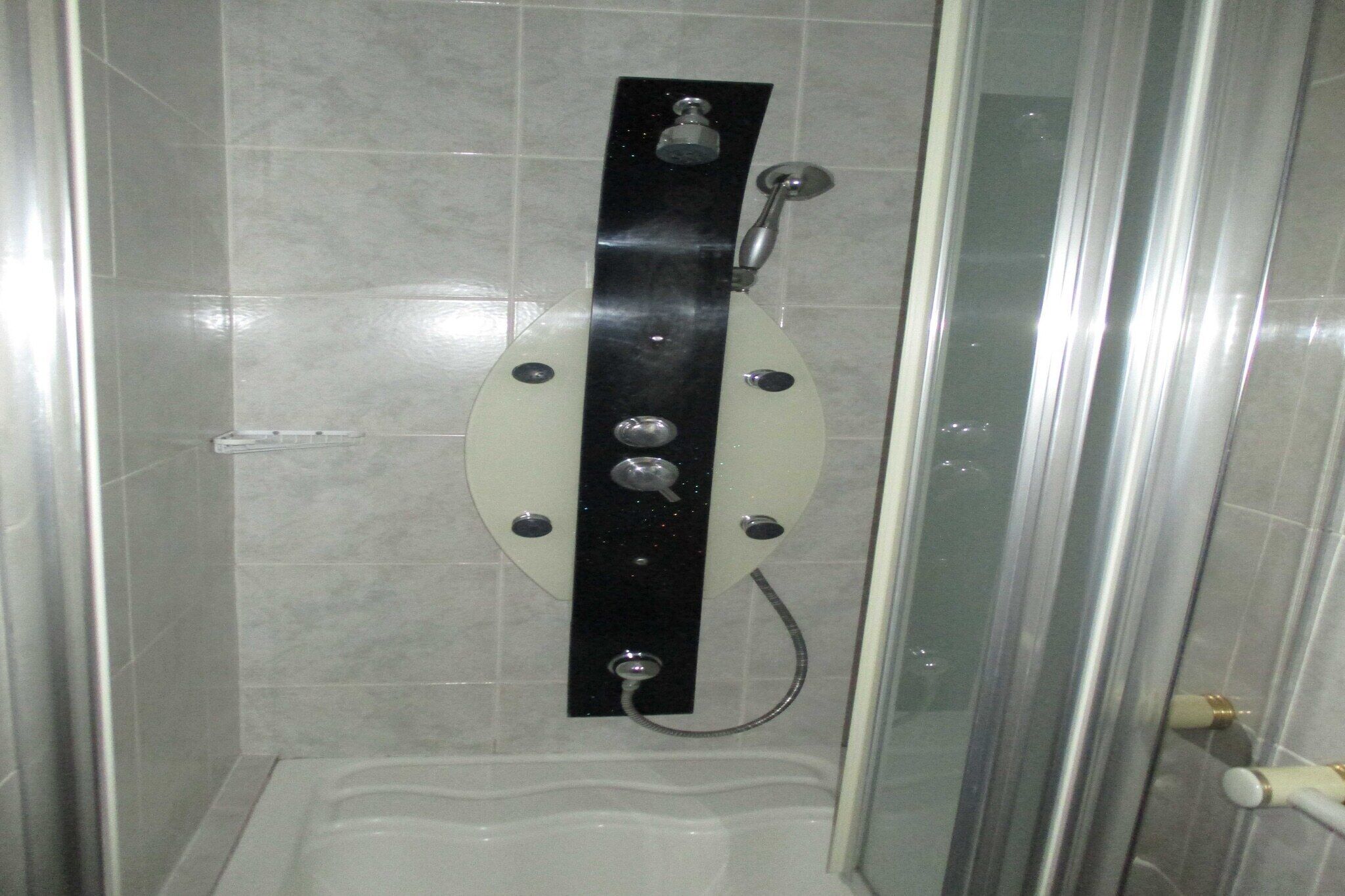 A glass-enclosed shower stall with a white floor, a black control panel, and a handheld showerhead.