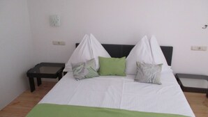 Ein ordentlich bezogenes Bett mit weißer Bettwäsche und grünen Akzenten, zwei Nachttische und eine schlichte Wand mit Lichtschalter.