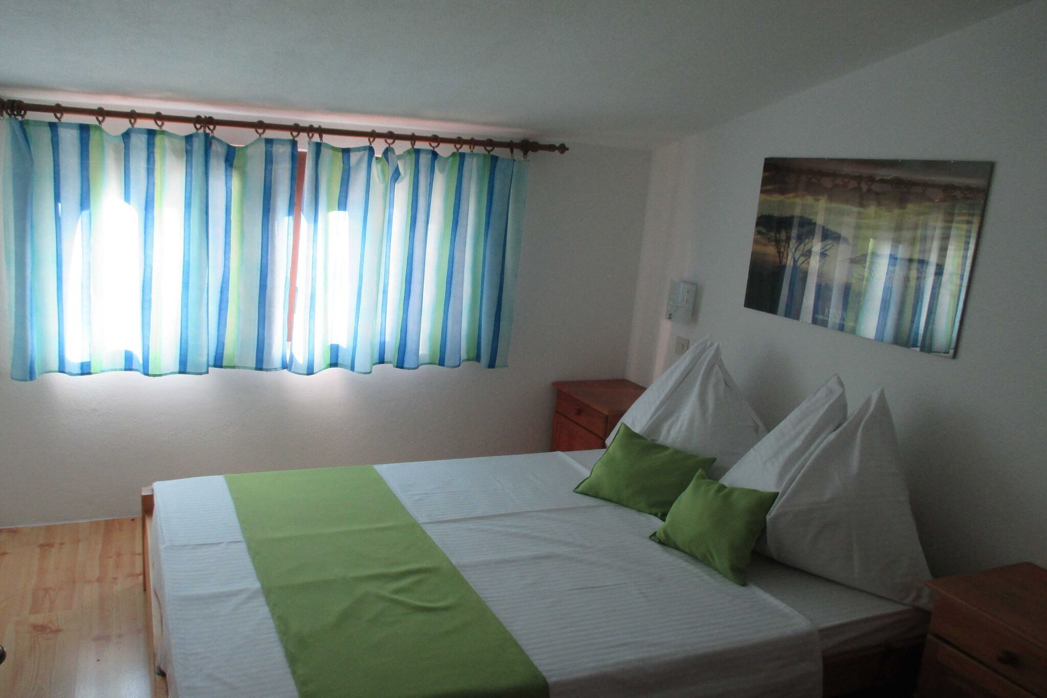 A bedroom with a bed, green pillows, a wooden nightstand, a white bedside table, and a window with blue curtains.