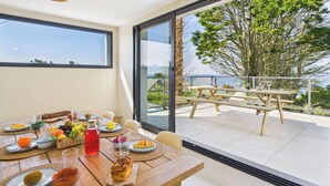 Un coin repas avec une table dressée pour le dîner, une bouteille de vin rouge et une vue sur la mer à travers de grandes fenêtres.