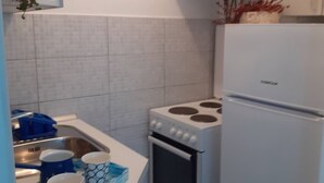 Una cocina con estufa, horno y refrigerador blancos. Se ve un escurreplatos azul con tazas y un lavamanos con grifo.