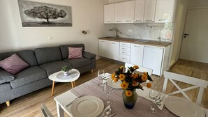 Een moderne woonkamer met een grijze bank, een klein rond tafeltje met een bloevas, en een eethoek voor twee personen met witte borden en glazen.