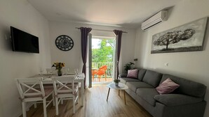 Een woonkamer met een grijze bank, een eettafel met stoelen, een aan de muur bevestigde televisie en een raam met gordijnen.