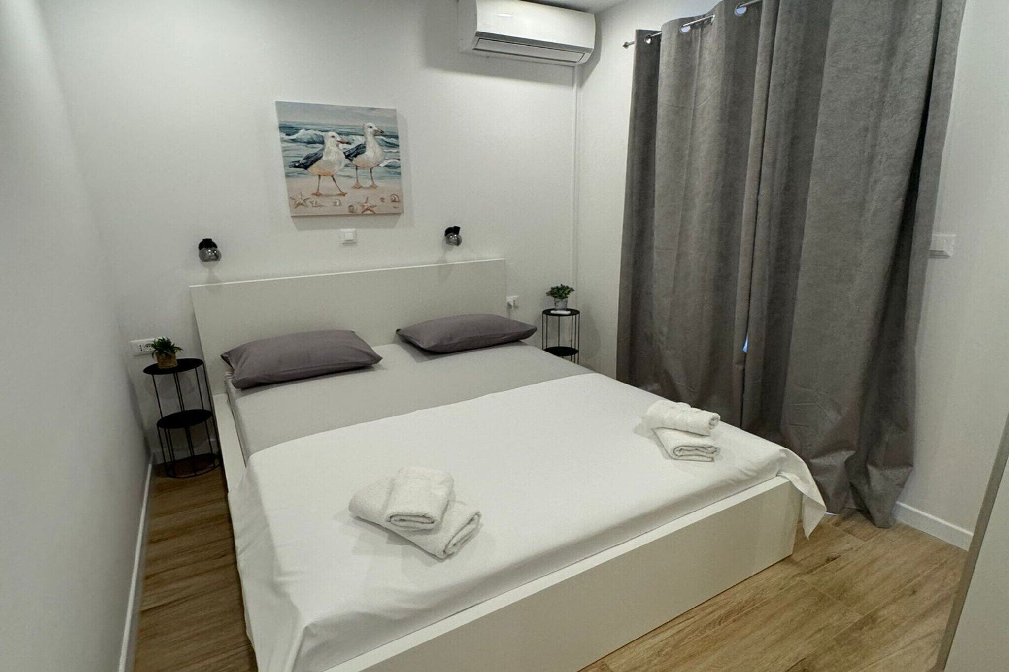 Una cama bien hecha con sábanas blancas y dos almohadas, una mesita de noche con una planta, y un cuadro de gaviotas en la pared.