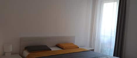 Una cama individual con cabecera, dos almohadas, una mesita de noche con lámpara, y una ventana con cortinas traslúcidas.