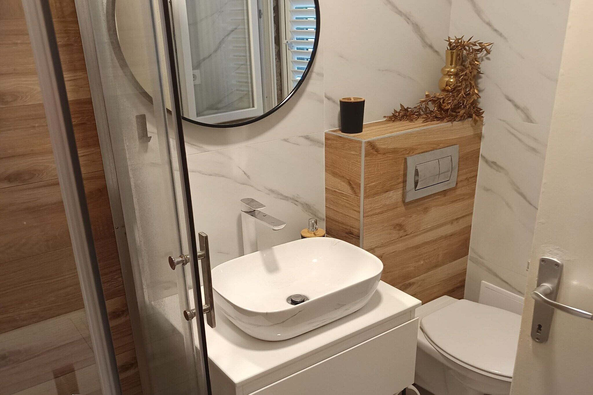 Et badeværelse med hvid vask, toilet, spejl og et træskab med lyskontakt.