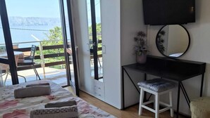 Ett sovrum med en säng, ett skrivbord, en stol, en tv och en balkong med utsikt över havet.
