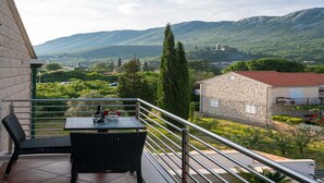 A balcony with wicker furniture and a table, overlooking a scenic view of mountains and a stone building.