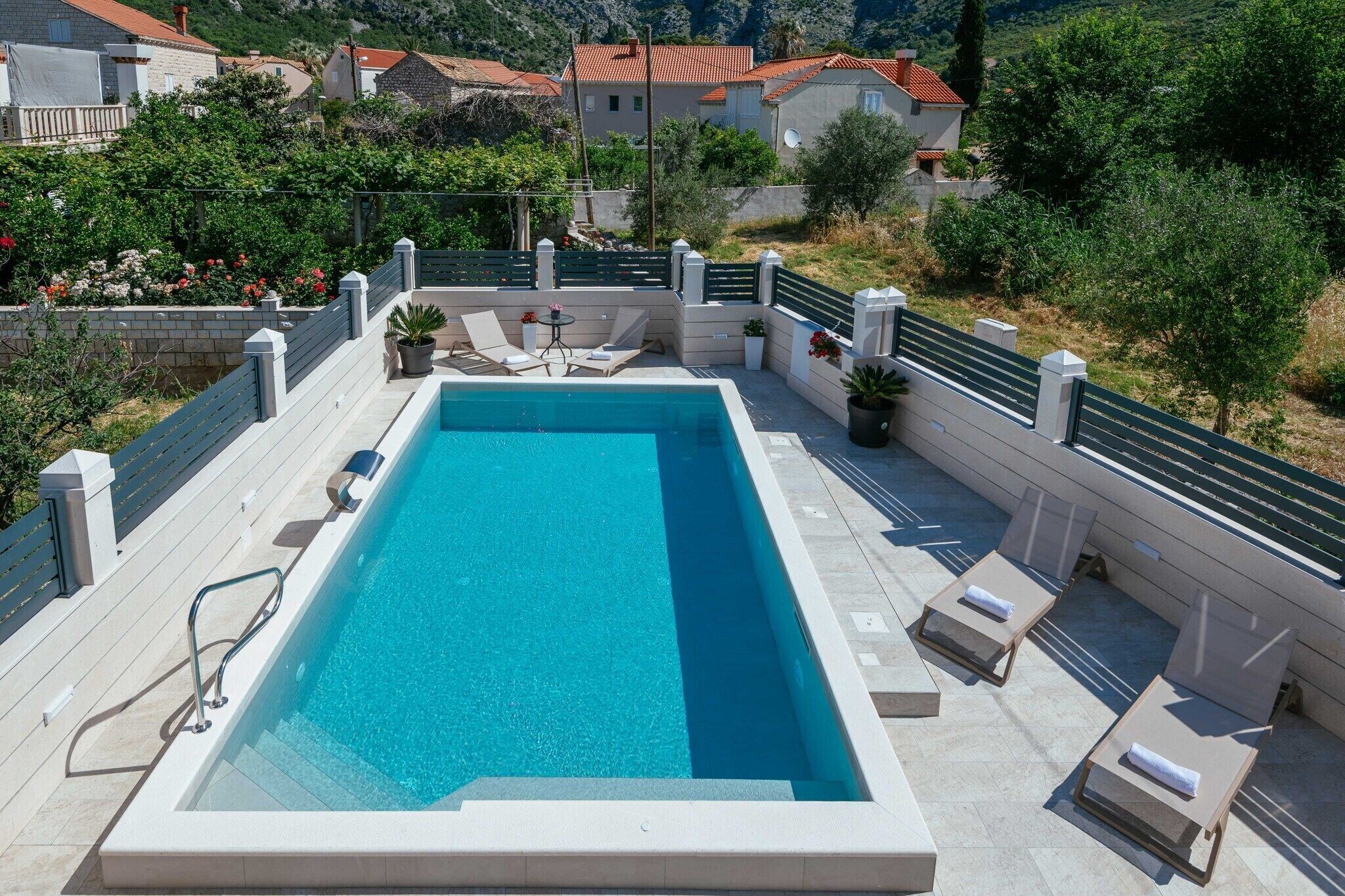 A rectangular swimming pool with a clear blue water, surrounded by a white deck and a green fence. There are lounge chairs and a small table with a potted plant.