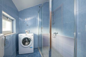 Une petite salle de bain avec des carreaux bleus, un lave-linge, une cabine de douche en verre et une fenêtre.