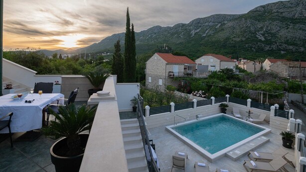 A rooftop pool area with a view of mountains, a dining table with chairs, and a small table with a glass of wine.
