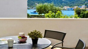 Ein runder Metalltisch mit Obstschale, Blumentopf und weißer Tasse auf einem Balkon mit Blick auf ein Gewässer und eine Hügelkuppe mit Gebäuden.