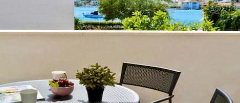 A round metal table with a bowl of fruit, a potted plant, and a white cup on a balcony overlooking a body of water and a hillside with buildings.