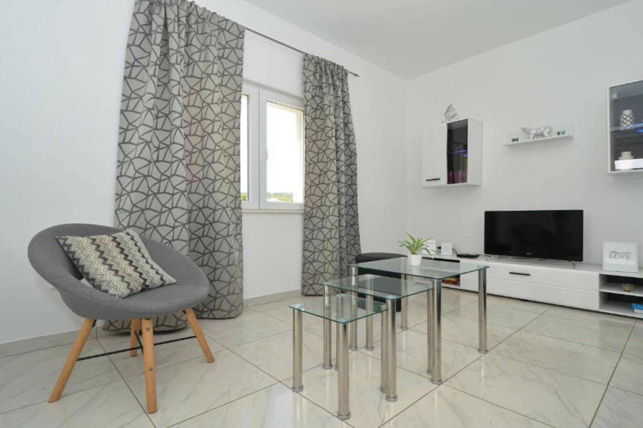 A modern living room with a glass coffee table, a grey armchair, a flat-screen TV, and a patterned curtain.