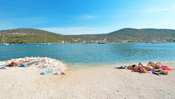 Ein Strand mit badenden Personen, einem klaren blauen Meer und Hügeln im Hintergrund.