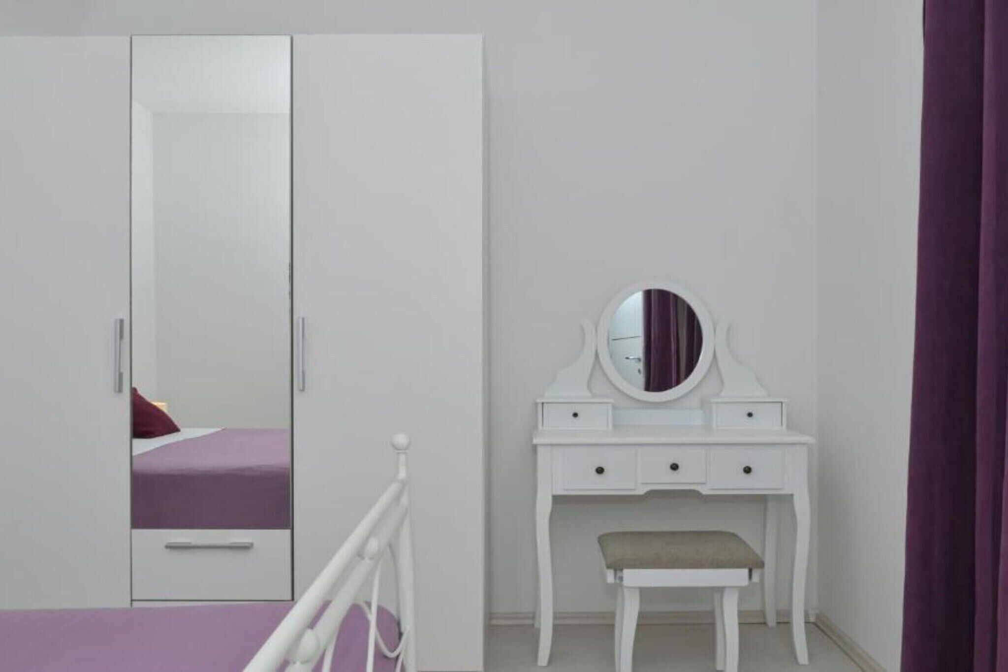 A bedroom with a white wardrobe, a bed with a purple bedspread, a white vanity with a mirror, and a bench.