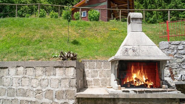 Una chimenea de piedra al aire libre con una chimada, rodeada por un muro de piedra y una zona ajardinada con una estructura de madera al fondo.