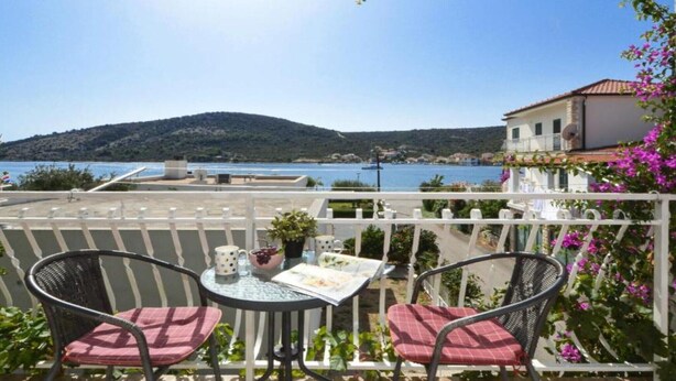 Un balcon avec deux fauteuils en osier, une petite table ronde et une vue sur une étendue d’eau et des collines en arrière-plan.