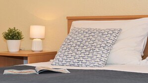 Un lit bien fait, agrémenté d’un coussin à motifs, d’une lampe de chevet et d’une plante en pot posée sur une table de chevet en bois.