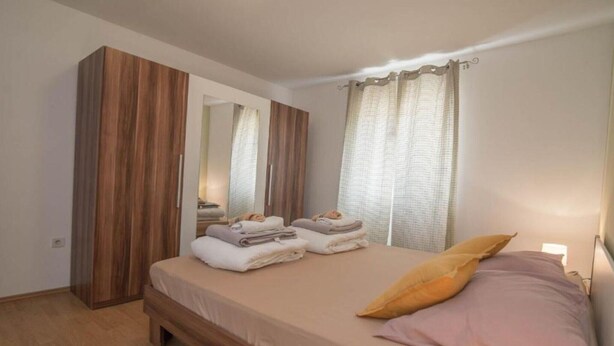 A bedroom with a bed, a wooden wardrobe, a mirror, and a window with curtains. - Apartments Bonic - Two Bedroom Apartment (Veli Losinj)