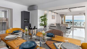 A modern dining area with a wooden table set for six, a kitchen area with dark cabinets, and a view of the sea through large windows. - Apartments Bregha - Three Bedroom Apartment with Terrace (Matulji)