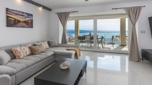 Een moderne woonkamer met een grijze modulaire bank, een zwarte salontafel en een groot raam met uitzicht op zee.