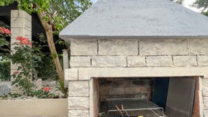 Un barbecue extérieur en pierre, doté d’une grille en métal, entouré d’un jardin avec des fleurs rouges et d’un arbre.