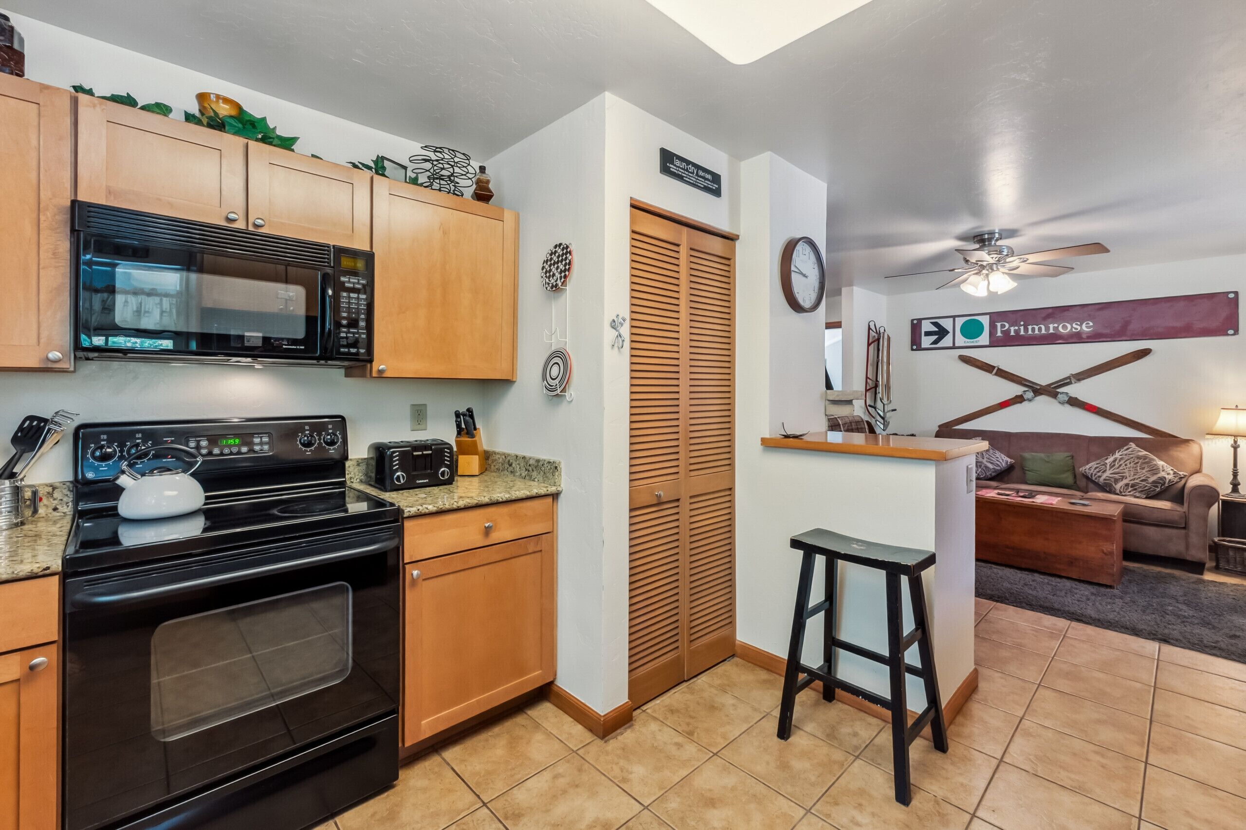 A kitchen with wooden cabinets, a microwave, an oven, and a coffee maker. There is a small island with stools and a sign that says "Primrose."