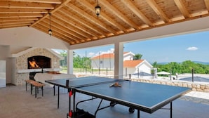 Un espace extérieur aménagé avec une table de ping-pong, une cheminée en pierre et une vue sur les maisons et la verdure.