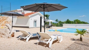 Un espace aménagé au bord de la piscine, avec des chaises longues blanches et des parasols, un mur en pierre et un ciel d’un bleu limpide.