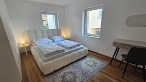A bedroom with a bed, bedside tables, a desk, and two windows.