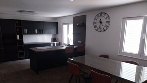 Una cocina moderna con armarios oscuros, una isla central y una zona de comedor con mesa de vidrio y sillas naranjas.