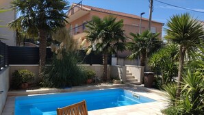 Una piscina con una silla de madera al frente, rodeada de palmeras y plantas en macetas.