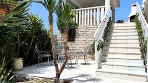 Una escalera conduce a un edificio blanco con un muro de piedra, rodeado de follaje y sillas de metal.