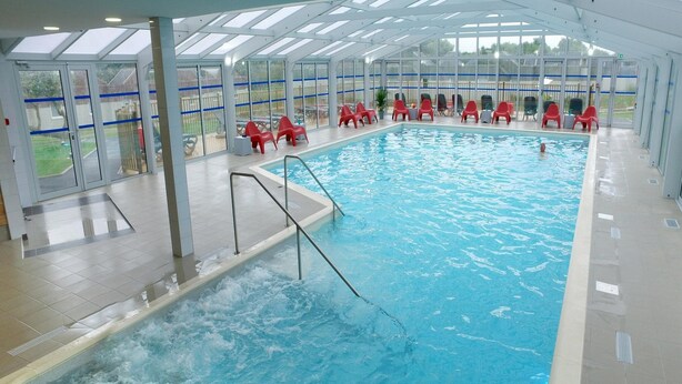 An indoor swimming pool with clear water, surrounded by red chairs and a glass enclosure with a view of the outside.