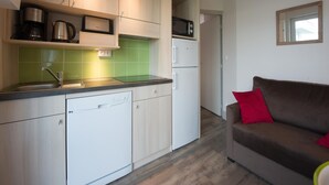 A compact kitchen with a dishwasher, a microwave, and a coffee maker. There is a brown sofa with a red pillow in the adjacent living area.