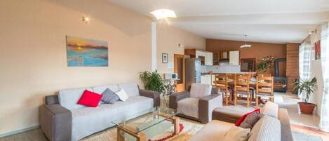 A modern living room with a sofa, armchairs, and a glass coffee table. There is a kitchen in the background with wooden cabinets and a dining area.