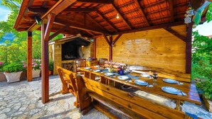 An outdoor wooden dining area with a long table set for a meal, benches, and a barbecue grill in the background.