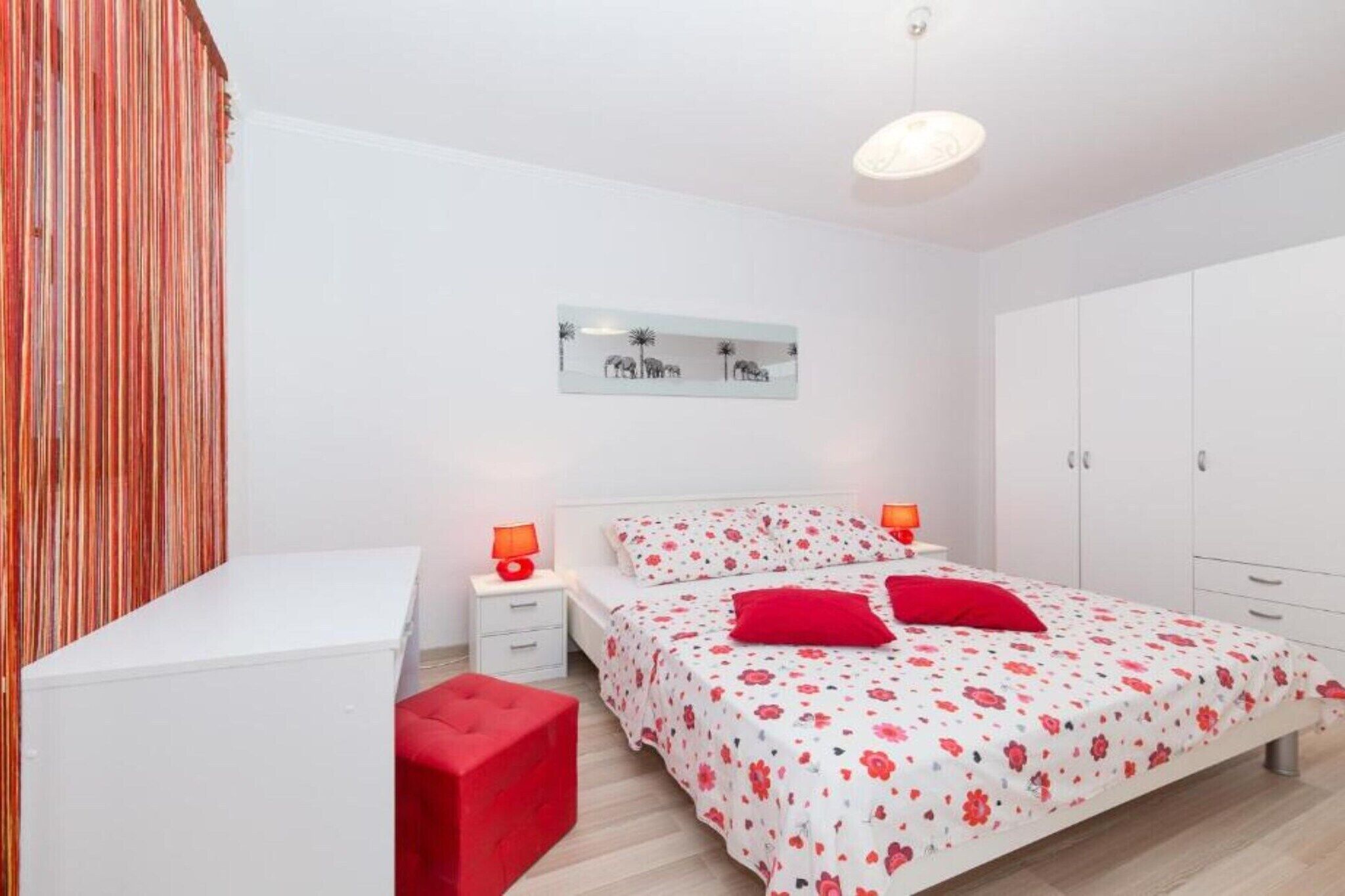 A bedroom with a bed, nightstands, a red bench, a wardrobe, and a wall-mounted artwork.