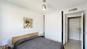 A bedroom with a bed, bedside tables, a wall-mounted lamp, a door, and a ceiling vent. - apartment near the beach (Fréjus)