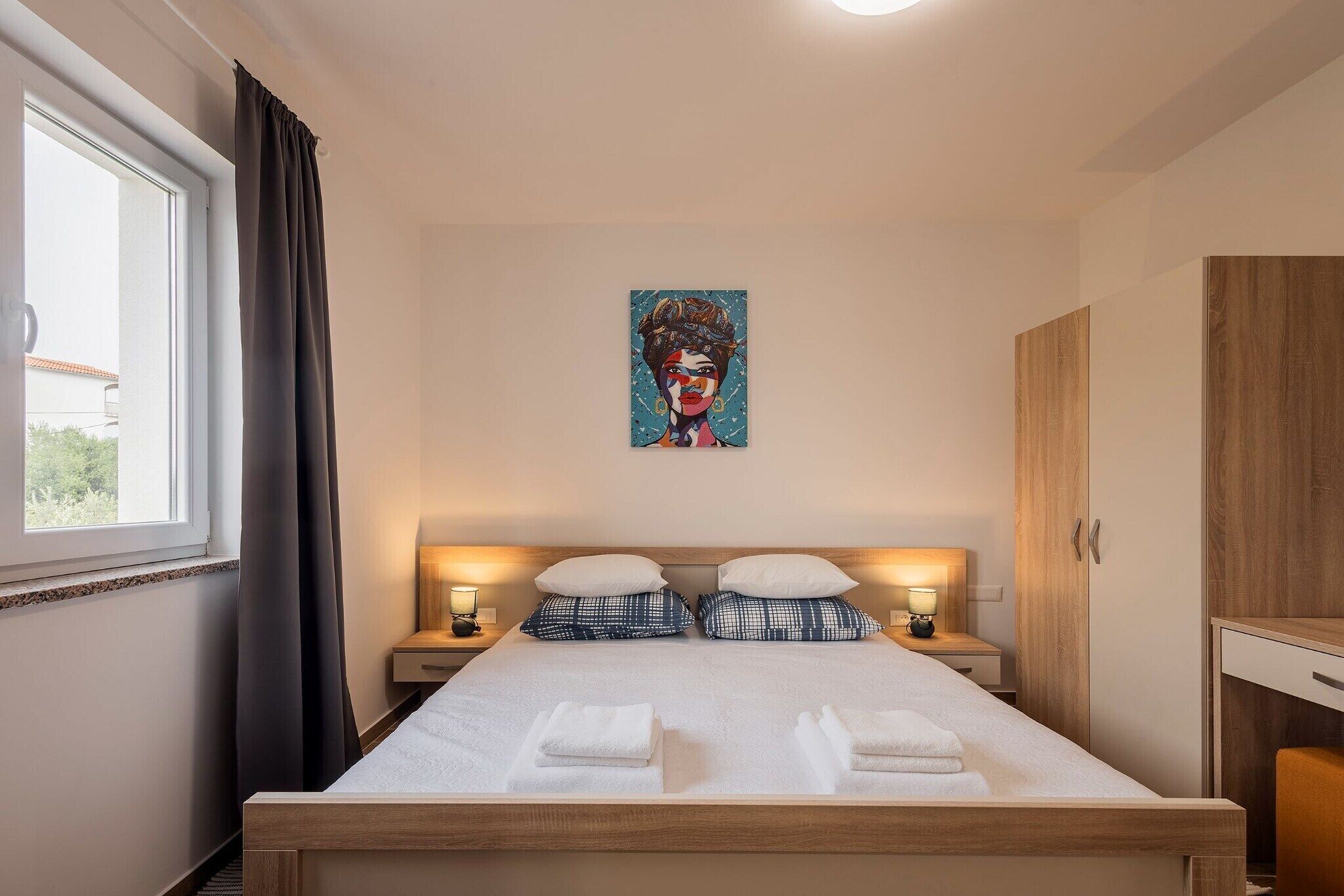 A neatly made bed with white linens and two pillows, a bedside table with a lamp, a painting on the wall, and a window with curtains.