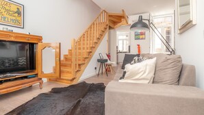 A modern living room with a wooden staircase, a flat-screen TV, a sofa, and a large rug.