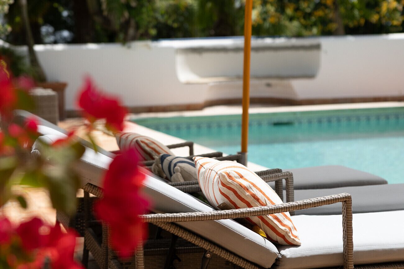 A poolside lounge area with wicker chairs and striped cushions, a pool, and a white wall in the background.