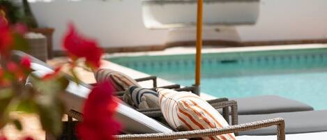 A poolside lounge area with wicker chairs and striped cushions, a pool, and a white wall in the background.