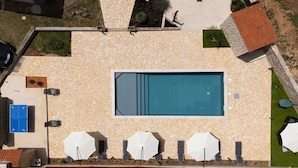 An aerial view of a swimming pool area with a stone deck, a rectangular pool, and several white umbrellas.