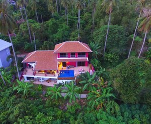 A two-story house with a red roof, surrounded by dense tropical vegetation. - PINK HOUSE (Koh Samui)