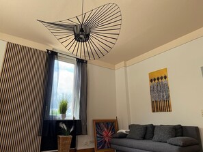 Una sala de estar con una lámpara de techo moderna, un sofá, una ventana con cortinas y una obra de arte en la pared.